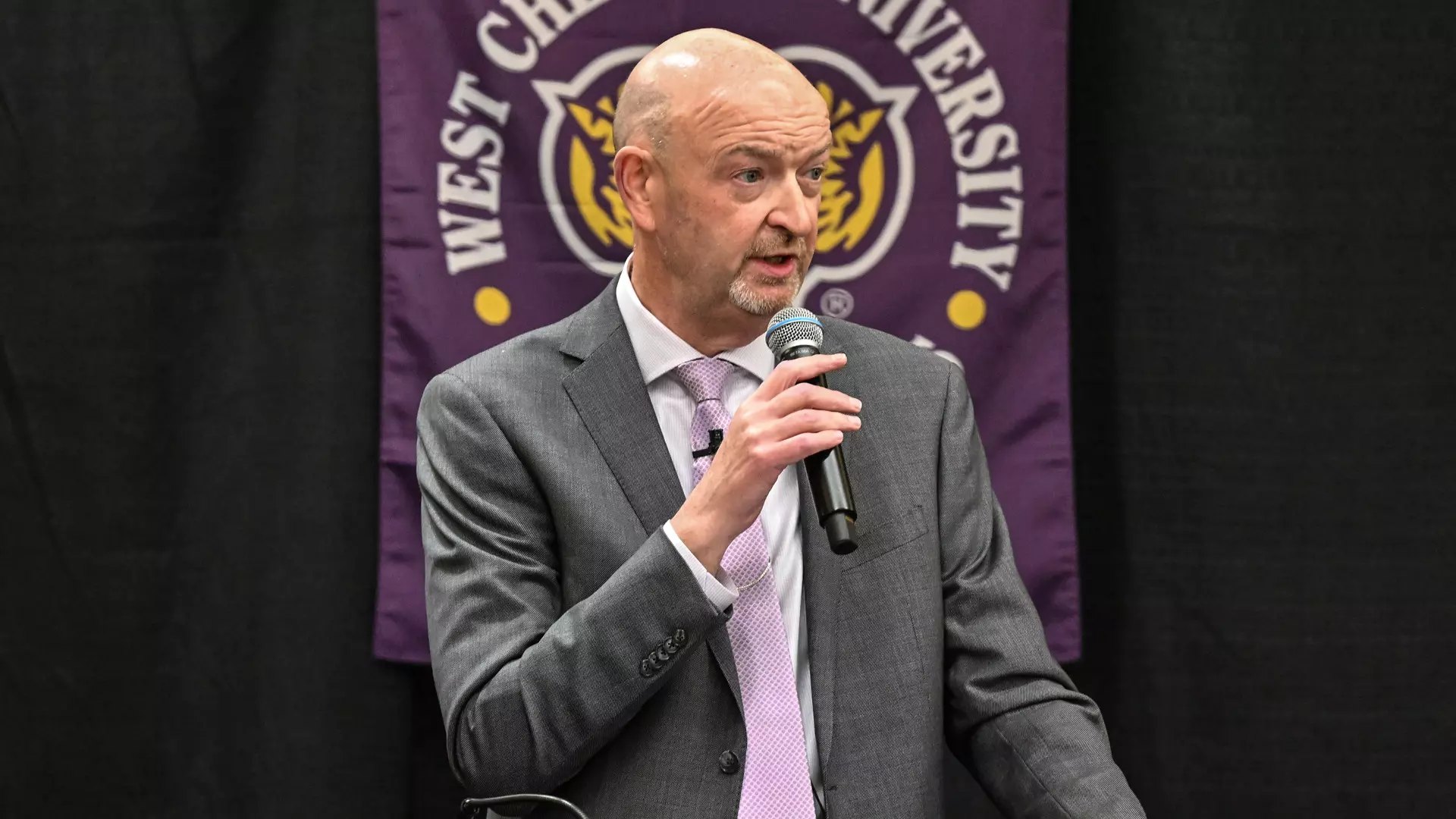 Man in gray suit and purple tie speaks into a handheld microphone before a purple university banner.