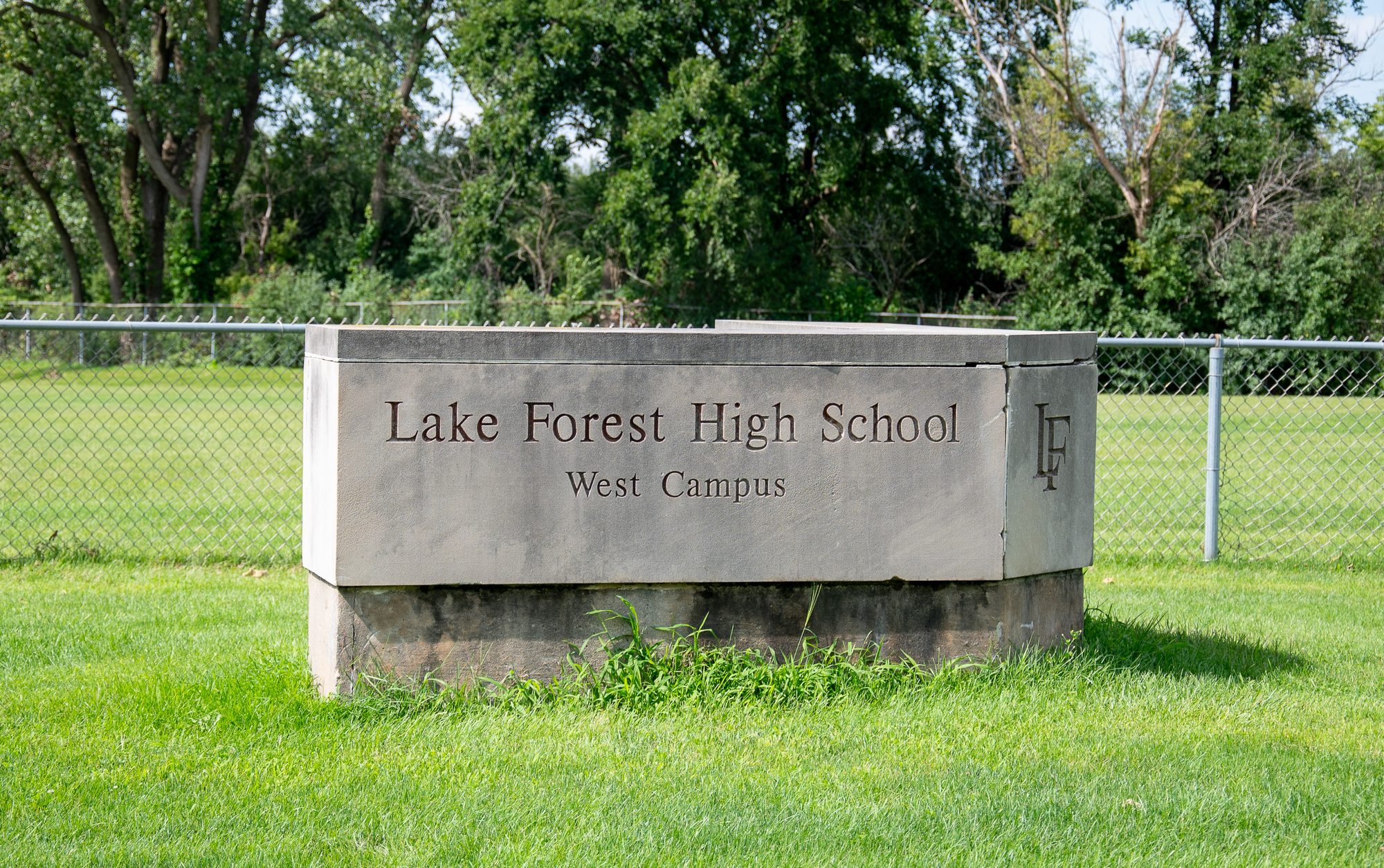 Stone entrance sign for a high school west campus on a green lawn beside a chain-link fence.
