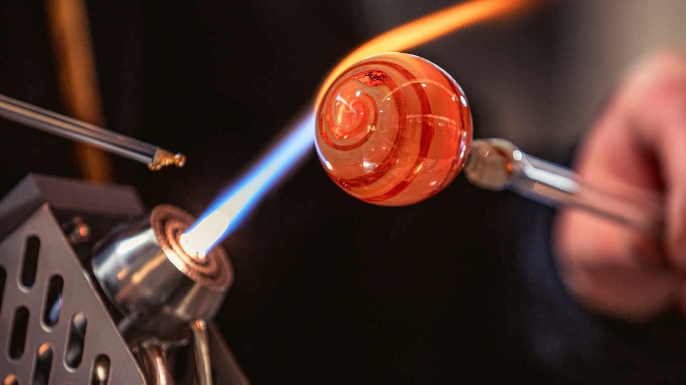 Close-up of a glassblower shaping a glowing orange glass sphere with a blue flame and metal tools.