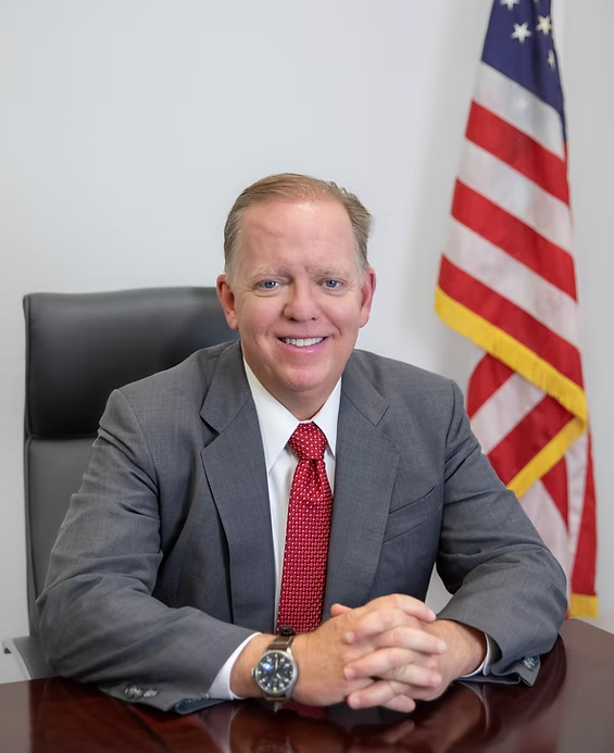 Herb Morgan seated at a desk in a gray suit and red tie, with an American flag behind him.