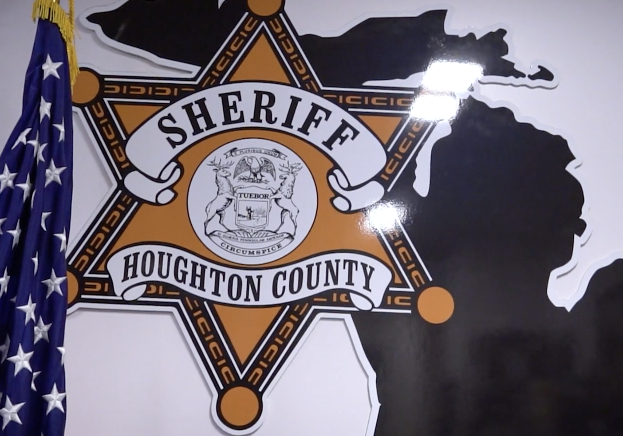 The Houghton County Sheriff's Office badge emblem mounted on a wall, next to an American flag.