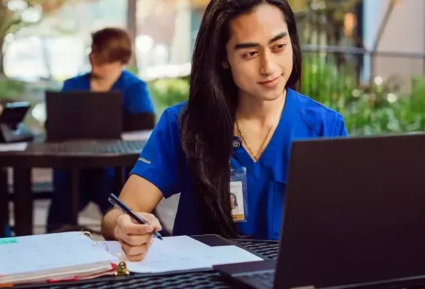 A Chamberlain University nursing student in blue scrubs writes in a binder while studying on a laptop at a table.