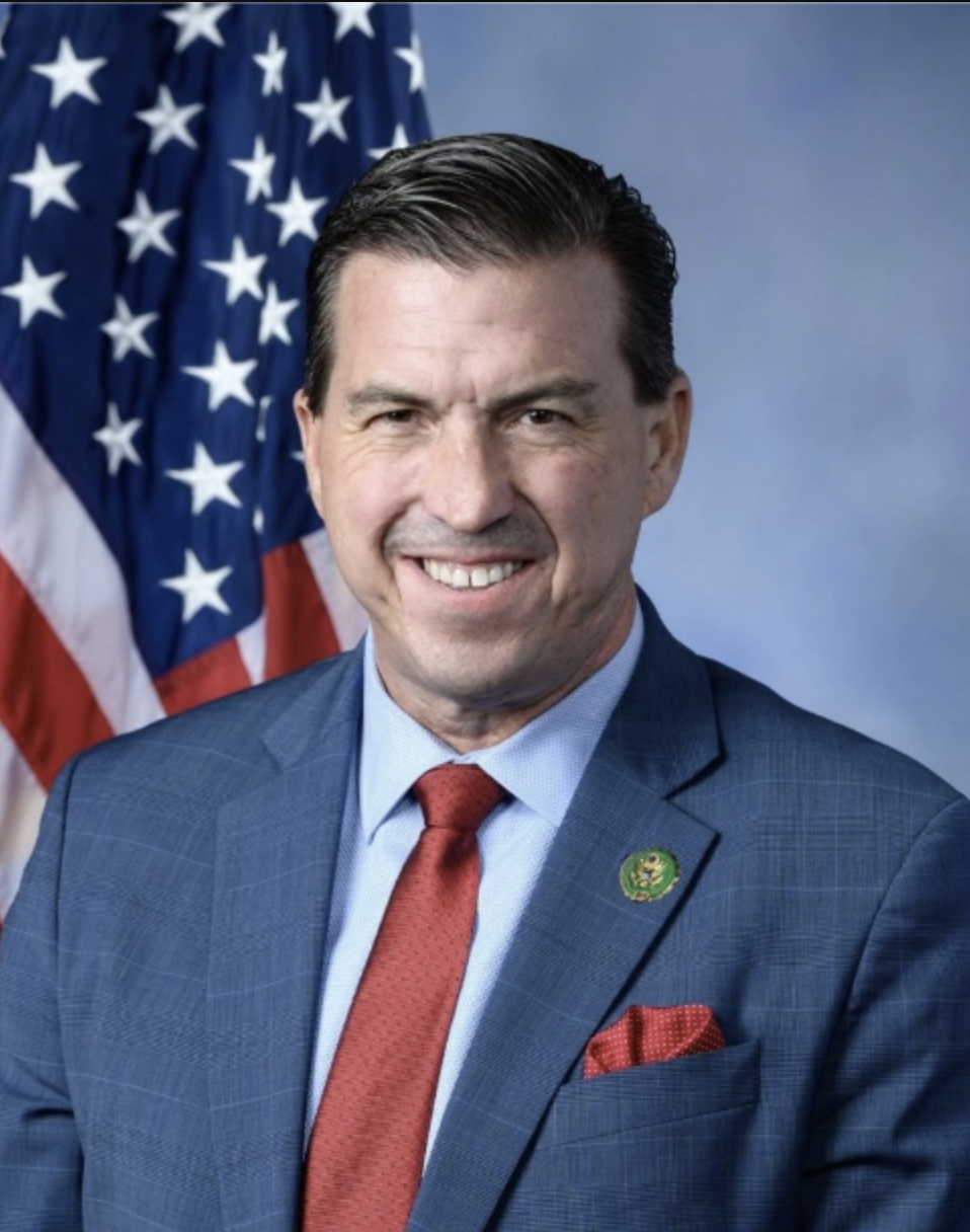 Official portrait of U.S. Rep. Kevin Mullin, wearing a navy suit and red tie, standing in front of an American flag.