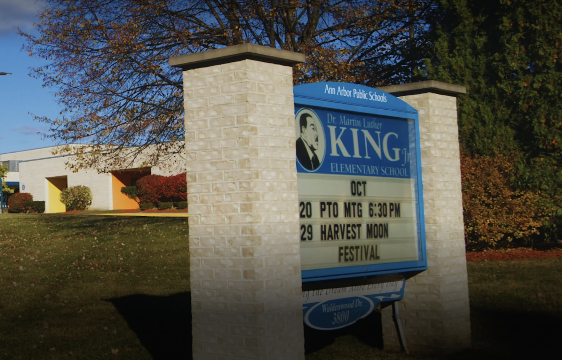 Blue King Elementary School sign between stone pillars, with the school building and autumn trees behind it.