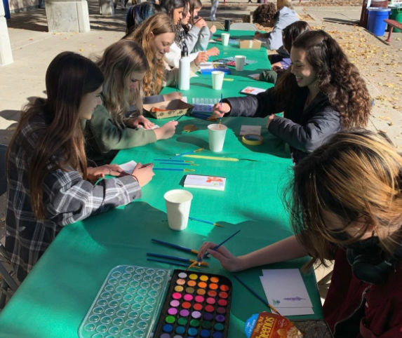 Teenage students seated at a long table covered with a green tablecloth, painting with watercolors outdoors on a school campus.