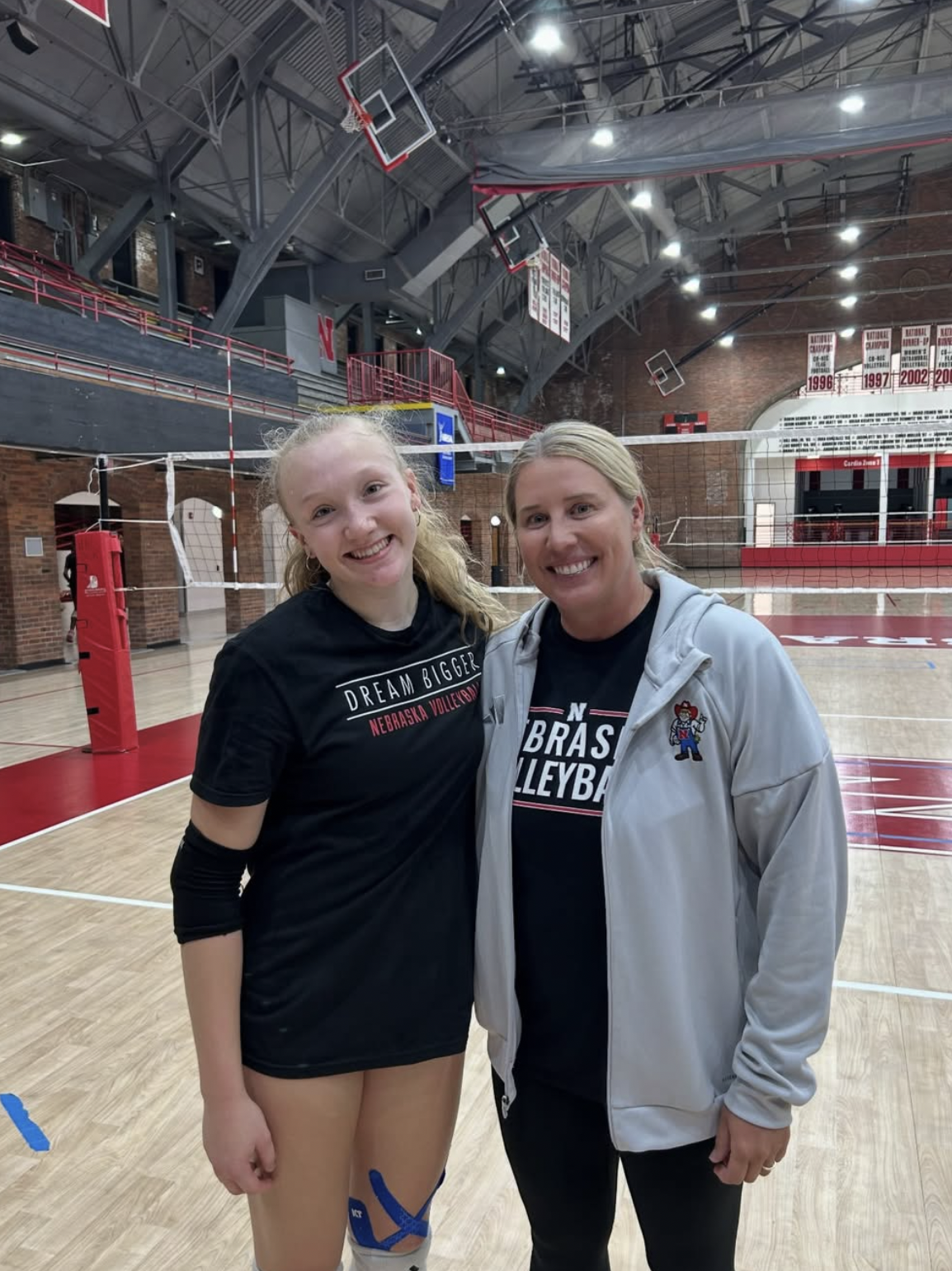 Northville volleyball player Lexi Coleman poses with Nebraska head coach Dani Busboom Kelly on an indoor volleyball court during the Huskers' Dream Team camp, with championship banners visible overhead.