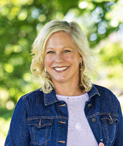 Lisa McIntyre smiles outdoors, wearing a denim jacket against a leafy background.