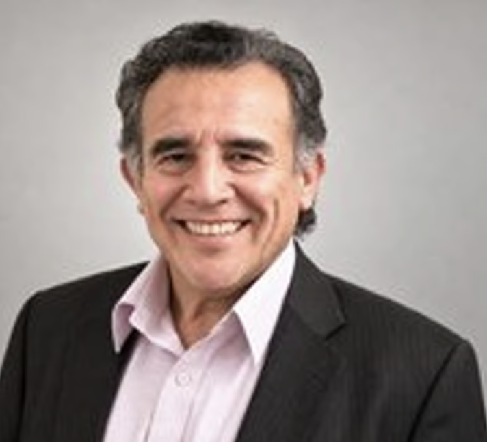 Luis Reynoso smiling in a dark jacket and light-colored shirt against a gray background.