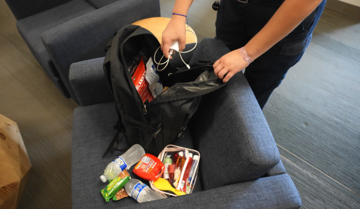 A person packs a black backpack emergency go bag with bottled water, snacks, a first aid kit and a phone charger.