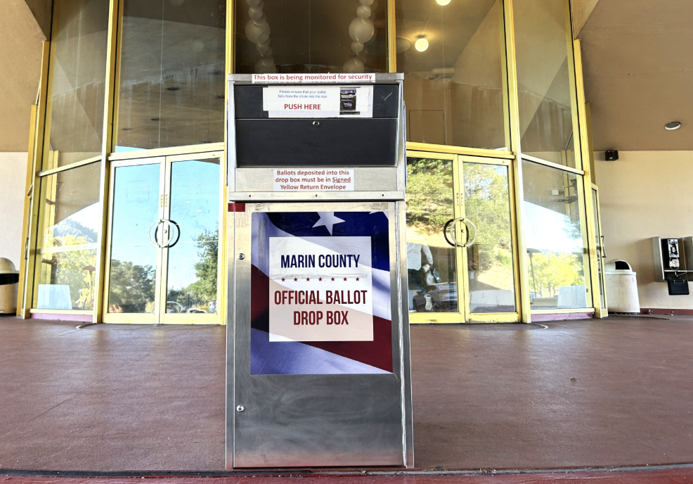 A stainless steel Marin County official ballot drop box stands in front of a glass building entrance.