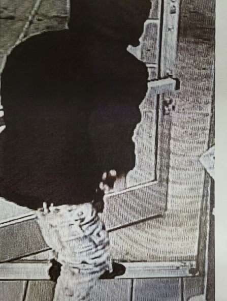 Grainy surveillance image showing a person in a dark hooded top and light-colored trousers near a doorway.