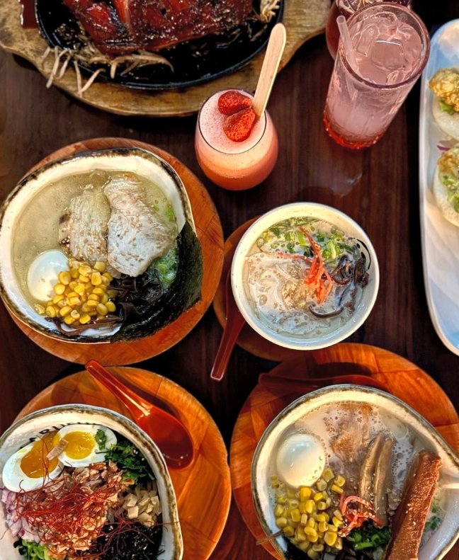 Overhead photograph of several ramen bowls, drinks and other dishes on a wooden table.