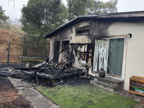 A burned exterior shed and scorched wall of a home, with a trampoline and play equipment in the yard, after a structure fire.