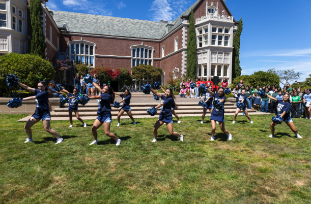 Mercy High School cheerleaders perform with blue pom-poms on the campus lawn as students watch behind them.