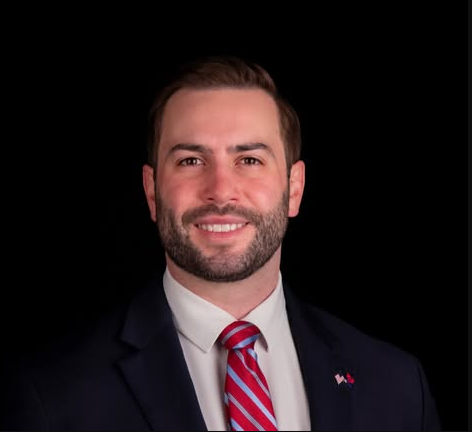Headshot of Michael Bouchard, Michigan congressional candidate, smiling in a navy suit with a red and white striped tie.