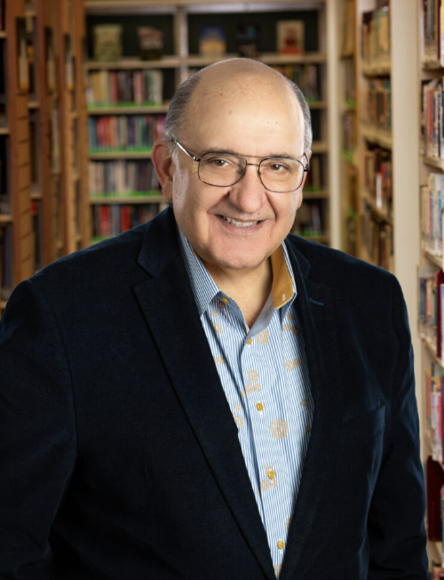 Michael Pappas smiles in front of library bookshelves, wearing glasses and a dark jacket.