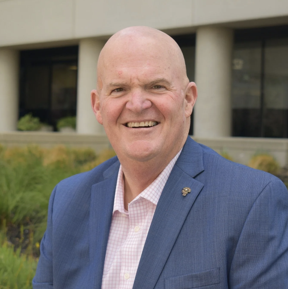 Mike Maxwell smiling outdoors in a blue jacket and pink checked shirt.