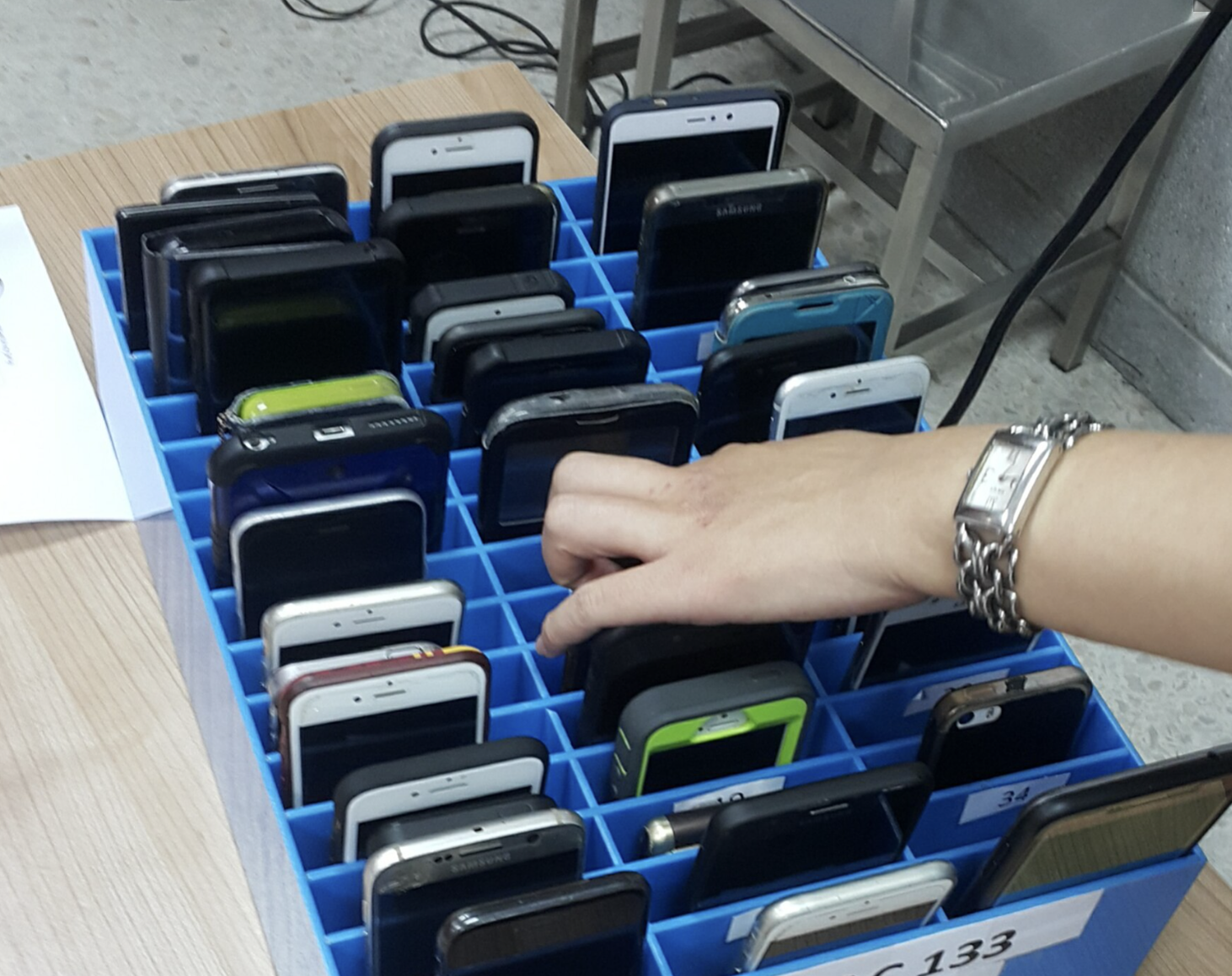 Rows of confiscated smartphones stored in slots of a blue plastic phone cage, with a hand reaching in to place a phone.