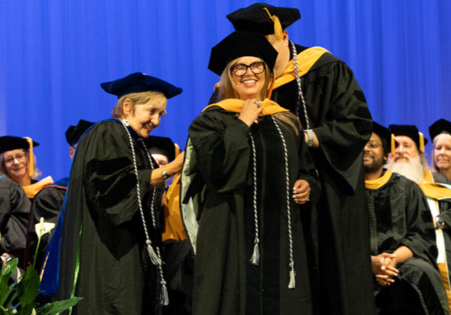 A Michigan School of Psychology doctoral graduate is hooded on stage during the 2026 commencement ceremony at The Hawk in Farmington Hills, smiling as faculty members assist with her academic regalia.