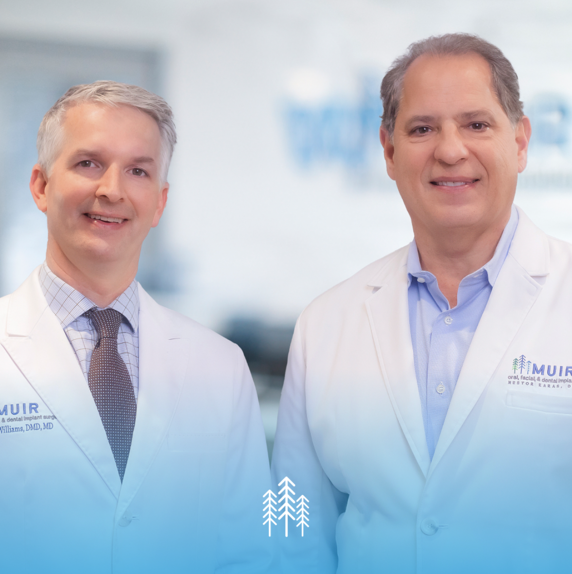 Dr. Brad Williams (left) and Dr. Nestor Karas (right) of Muir Oral, Facial & Dental Implant Surgery in white lab coats in their Walnut Creek office
