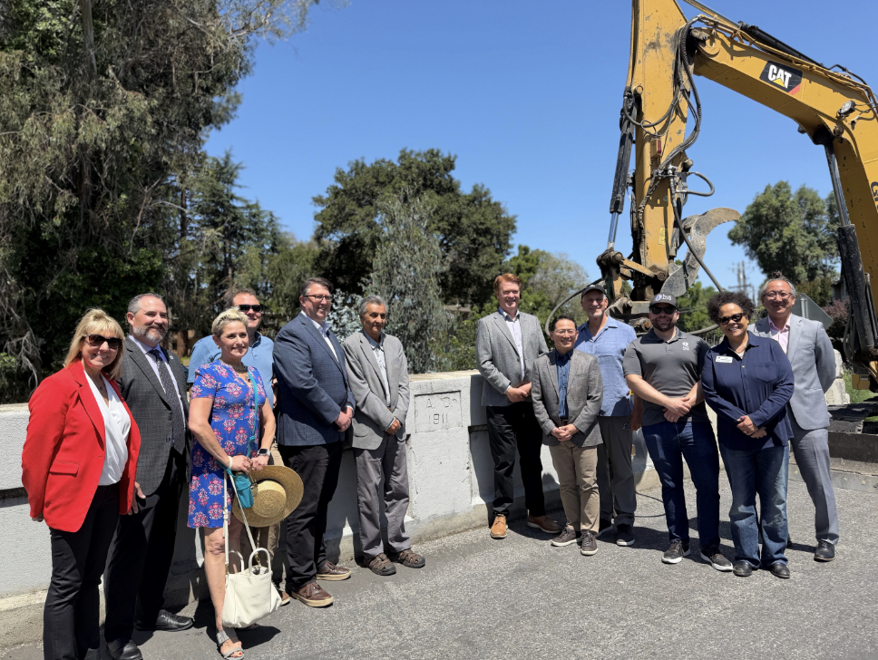 Palo Alto, East Palo Alto and Valley Water officials, including board member Rebecca Eisenberg, pose at the Newell Road Bridge groundbreaking with a construction excavator in the background