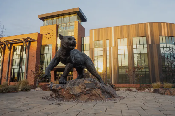 A bronze wildcat statue perches on a rock outside a brick and glass building on Northern Michigan University's Marquette campus at sunrise.