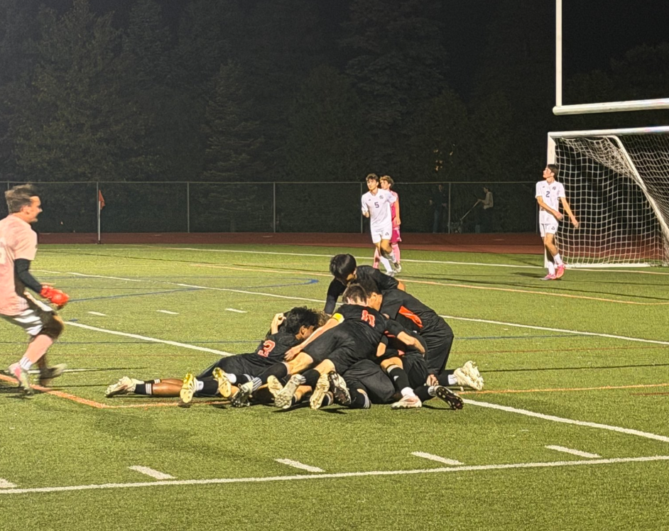 Soccer players in dark jerseys pile together in celebration on the field at night, as an opposing player in white looks on and a goalkeeper runs in from the sideline.