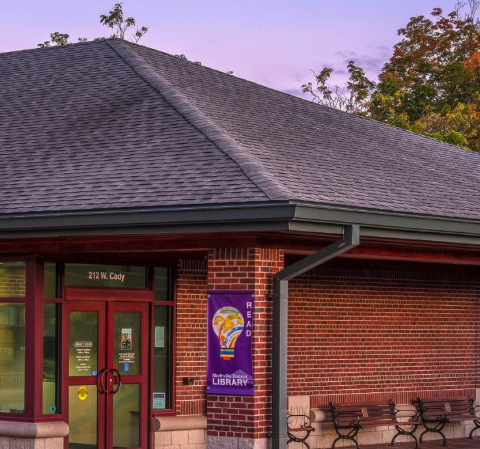 Brick exterior of Northville District Library, with red entrance doors, a purple library banner and benches.