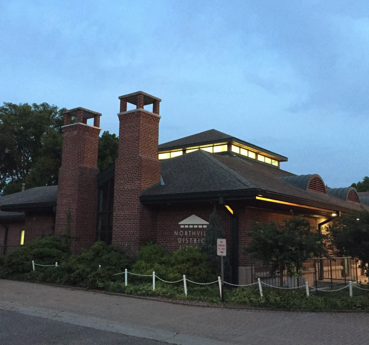 The Northville District Library building at dusk, its brick exterior and illuminated clerestory windows visible behind a low iron fence.
