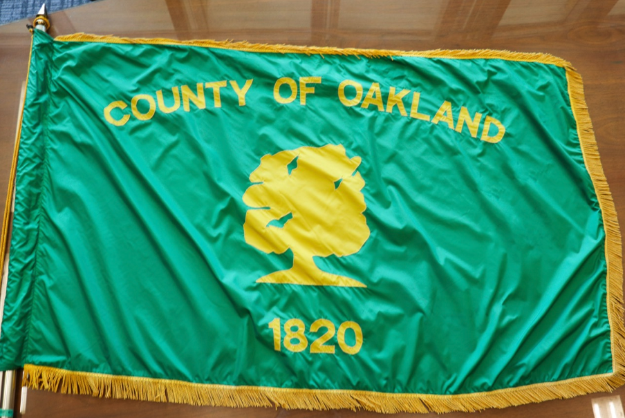 Green Oakland County flag with a yellow oak tree, the words “County of Oakland” and the number “1820.”