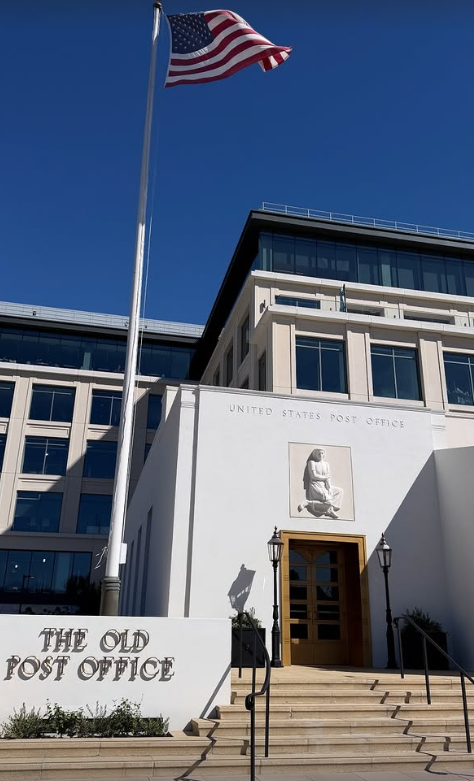 An American flag flies above The Old Post Office entrance, with a stone relief over the doors and a modern building behind it.