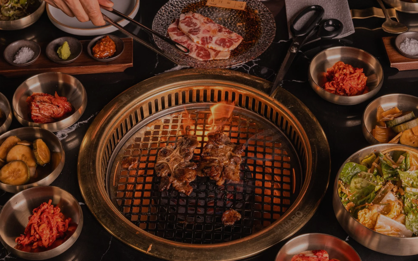A Korean barbecue table at OLHSO with meat grilling over an open-flame charcoal grill, surrounded by banchan side dishes like kimchi and pickled vegetables.