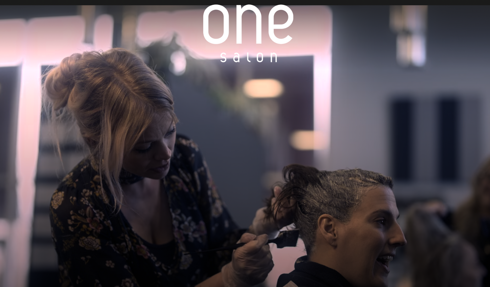 A One Salon stylist applies hair color to a client at the Novi salon, with the "One Salon" logo displayed above.