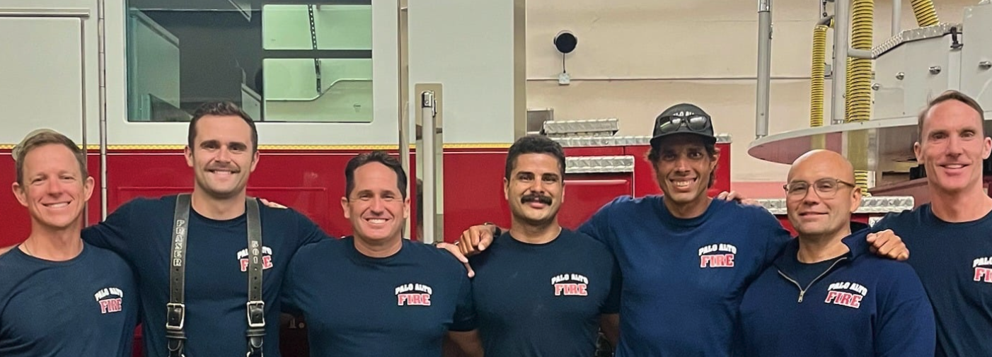 A group of seven men wearing navy "Palo Alto Fire" T-shirts pose together with their arms around each other in a fire station, standing in front of a red fire engine.