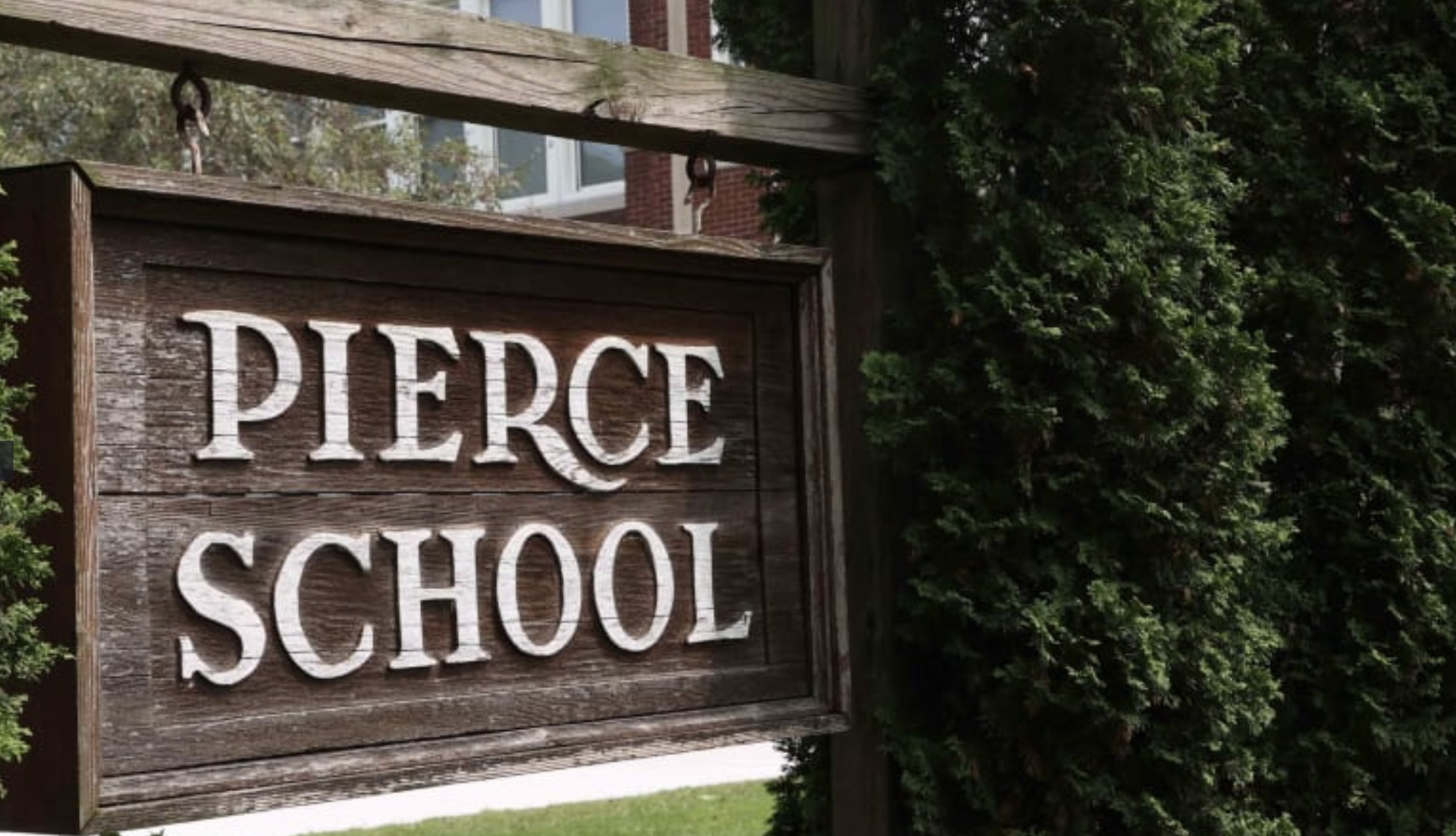 Wooden “Pierce School” sign beside evergreen shrubs, with part of the school building behind it.
