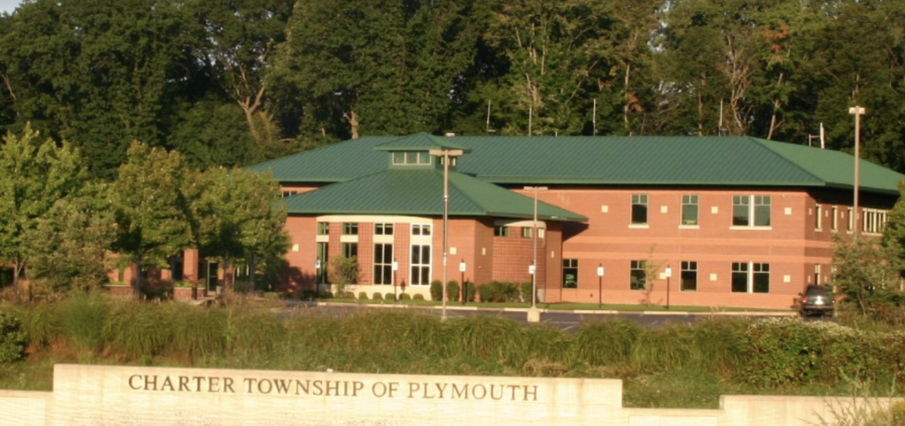 Exterior view of the Charter Township of Plymouth municipal building, a red-brick building with a green metal roof, with a stone sign reading "Charter Township of Plymouth" in the foreground.