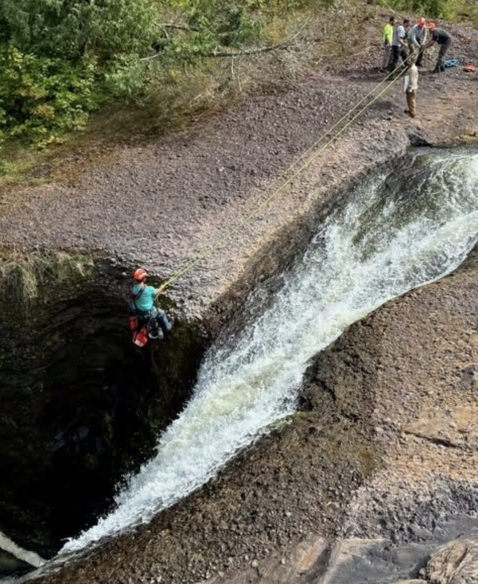 A rescuer rappels down the rock face beside Rainbow Falls during a rope rescue, with other responders anchoring the line at the top of the gorge.