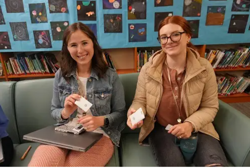 Two Rochester Community Schools staff members sit on a library couch holding their new Centegix CrisisAlert panic button badges.