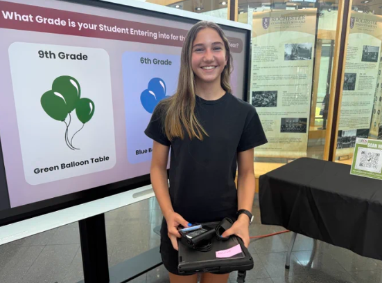 A Rochester Community Schools student poses with her new Chromebook at the district's device deployment event, with a sign directing 9th and 6th graders to separate check-in tables.