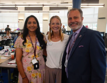 Three Rochester Community Schools staff members smiling together at a new-educator gathering, wearing lanyards, with a school library or learning space visible behind them.