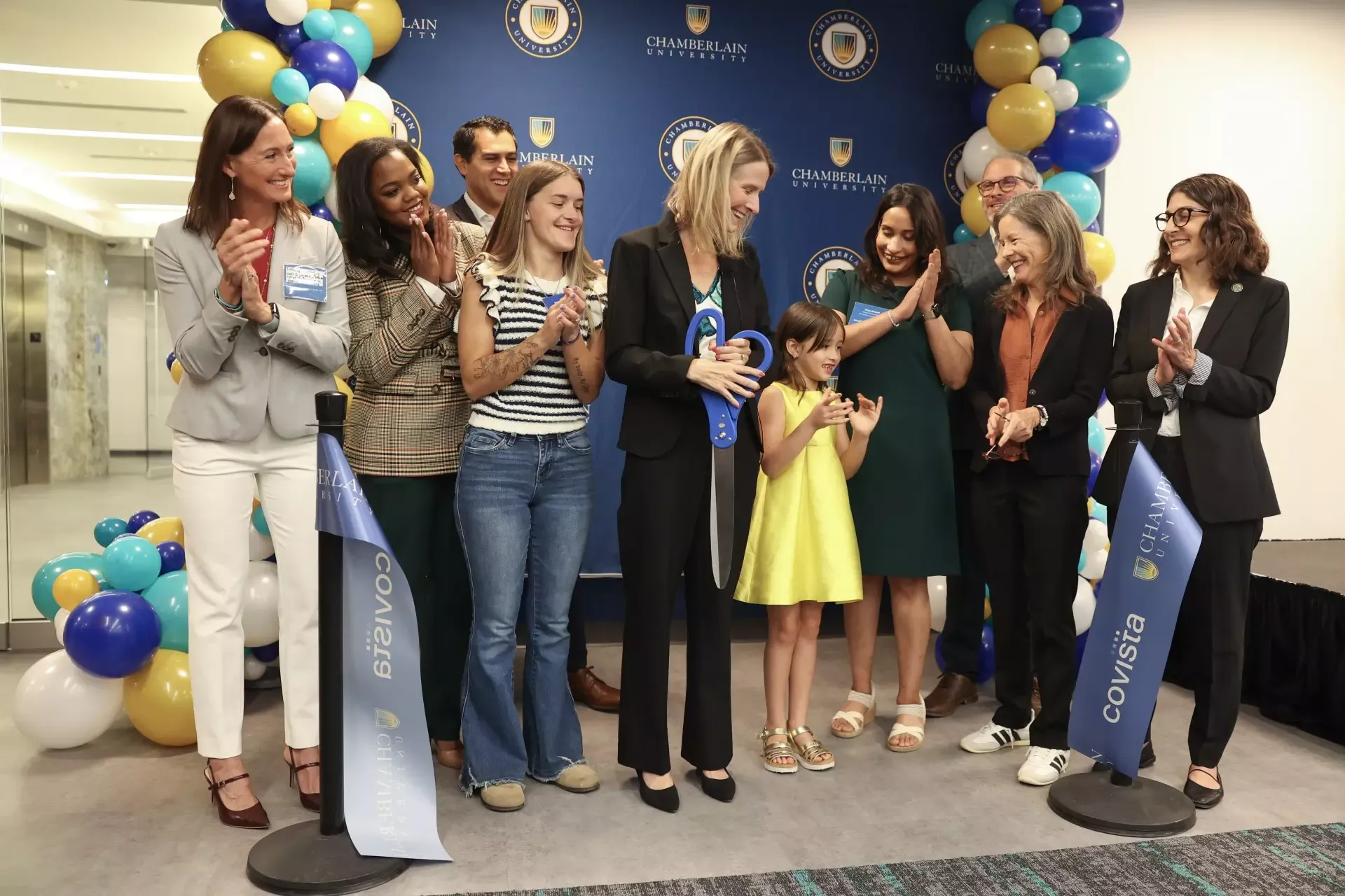 Attendees applaud as a woman holds oversized scissors at a Chamberlain University ribbon-cutting ceremony, framed by blue, teal, gold and white balloons.