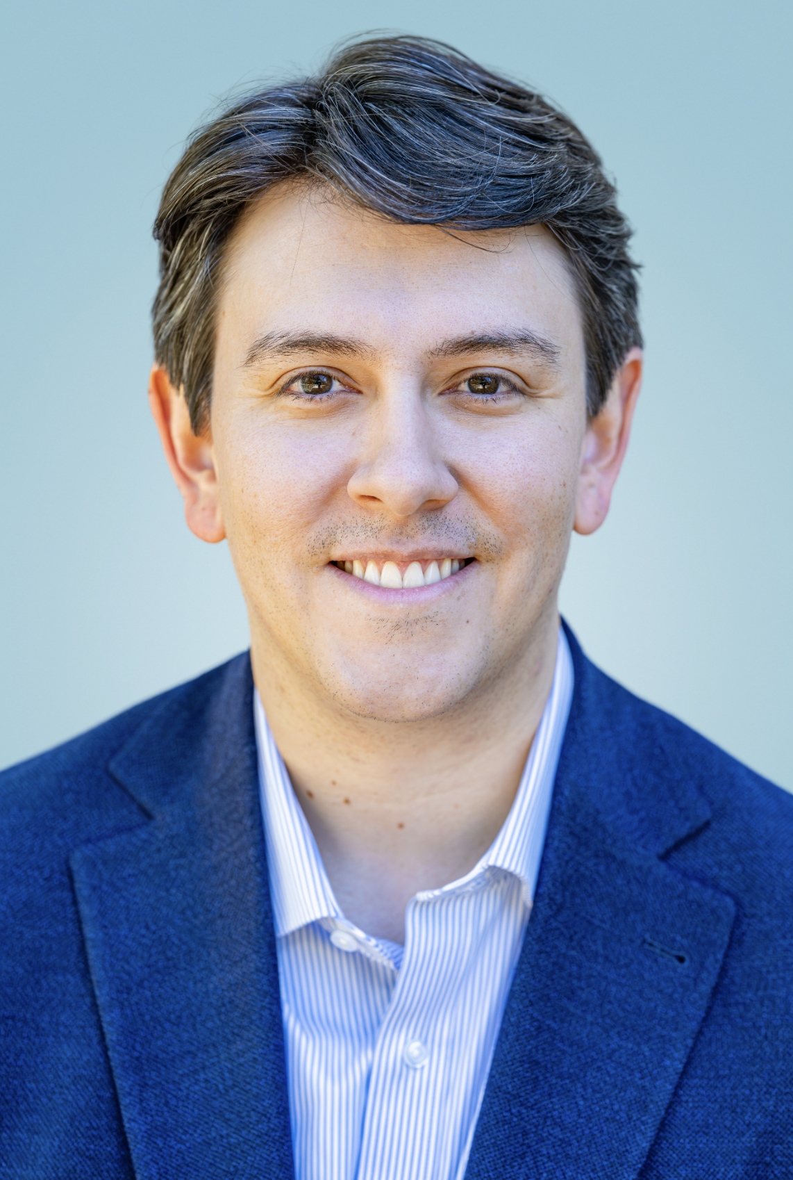 Robert Sandoval smiling in a blue blazer and striped shirt against a pale blue background.