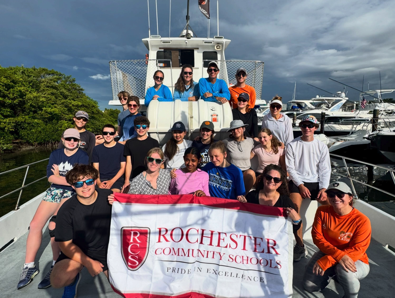 Rochester High School AP Capstone students and adults pose on a research boat while holding a Rochester Community Schools banner.