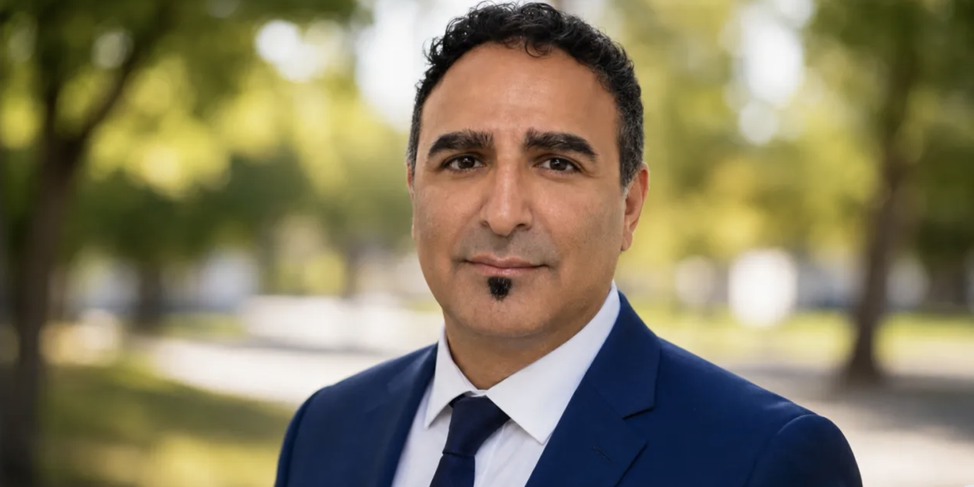 Rod Ehsani wearing a blue suit, white shirt and dark tie outdoors against a blurred green background.