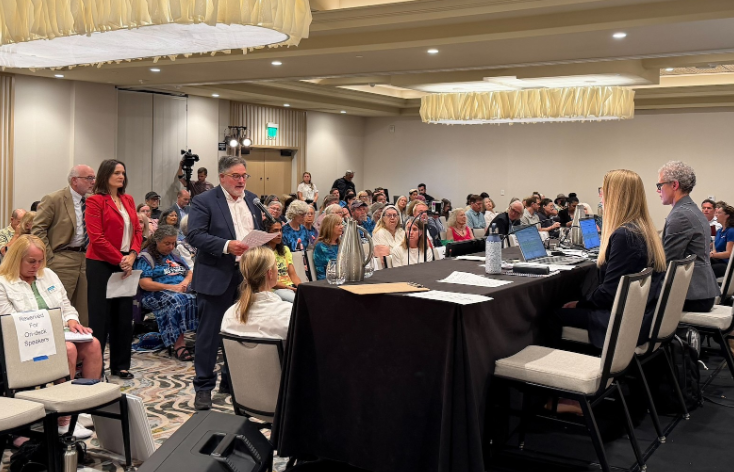 A man speaks at a podium before a panel at a crowded NOAA public hearing in Santa Monica, with rows of attendees seated behind him.