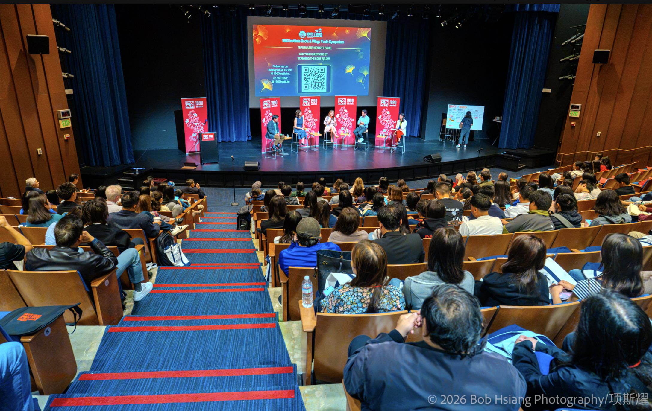 A full auditorium of attendees watches panelists speak on stage at the Roots & Wings Youth Symposium.