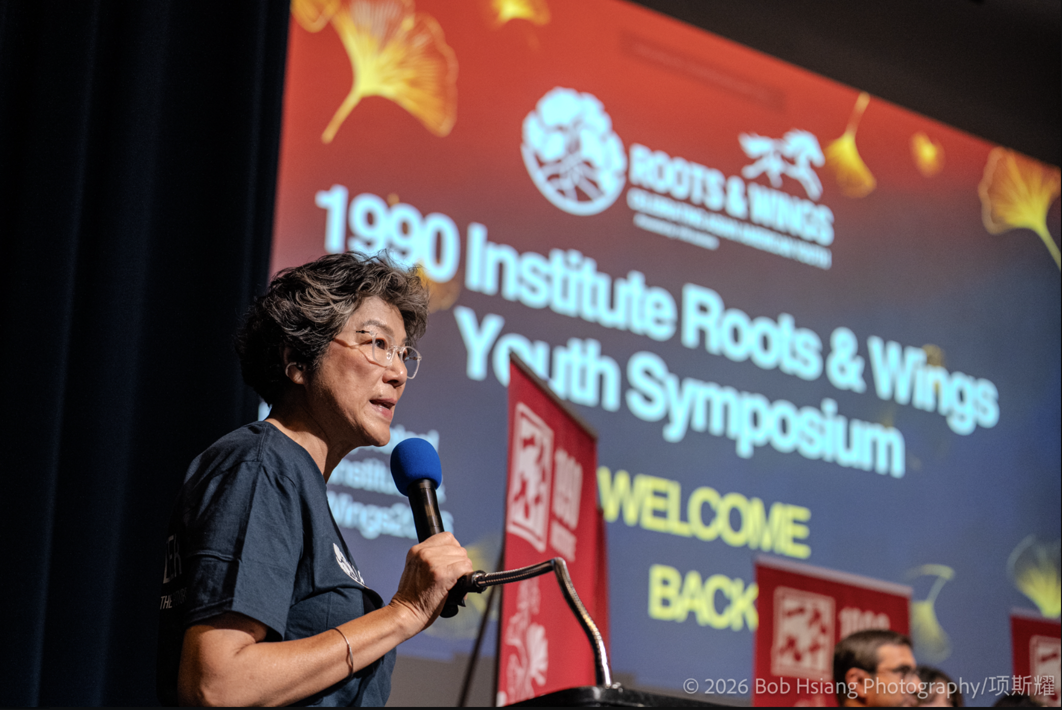 Sandra Pan speaks into a microphone on stage, welcoming attendees to the Roots & Wings Youth Symposium.