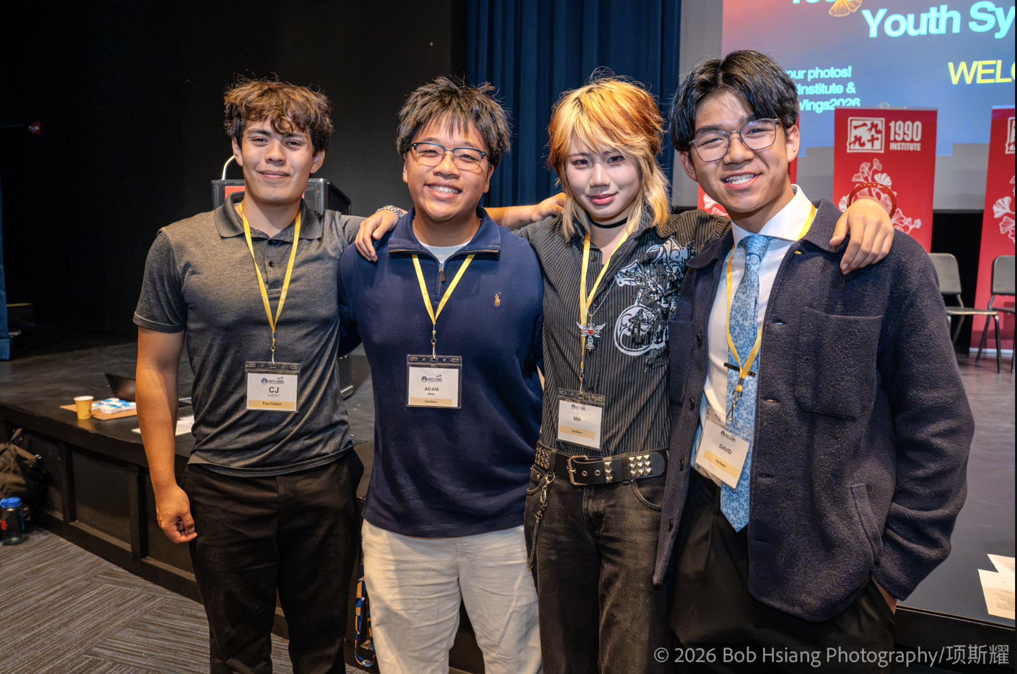Four AAPI student leaders stand arm in arm on stage, smiling, at the Roots & Wings Youth Symposium.