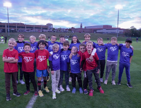 Youth soccer players from Rochester Soccer Club pose together in red and blue jerseys on a field at dusk.