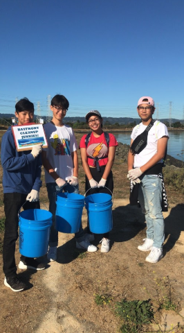 Four volunteers stand near the San Mateo bayfront holding blue cleanup buckets.